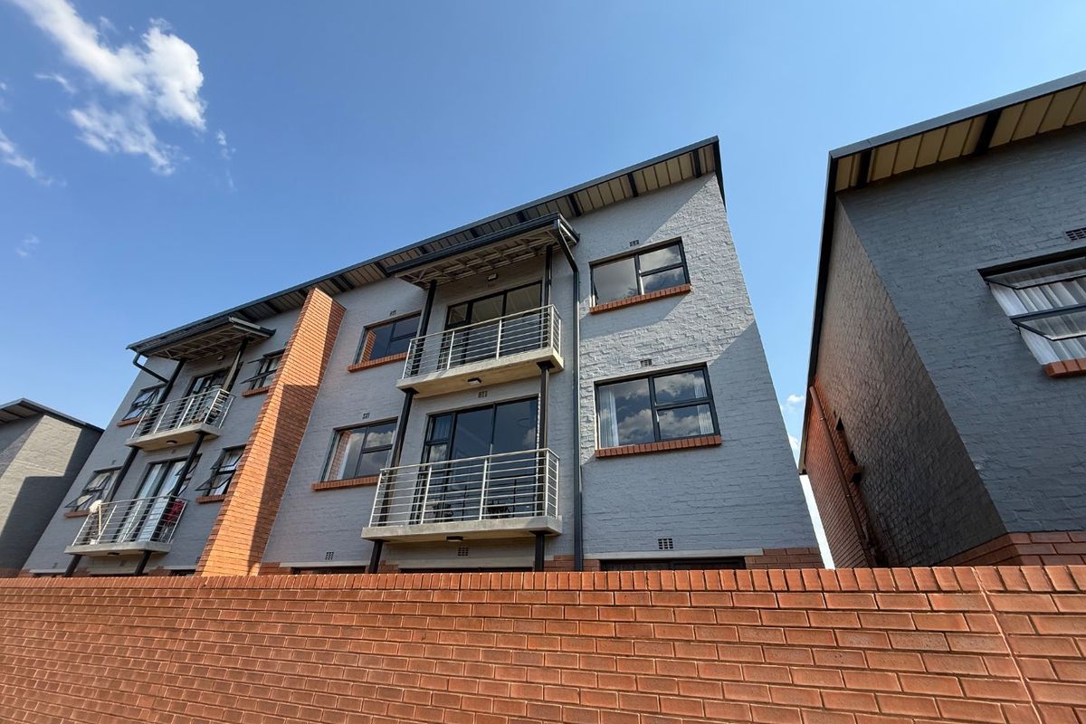 A low-angle exterior view of a modern apartment building featuring grey brickwork, multiple balconies with metal railings, and a prominent red brick boundary wall.