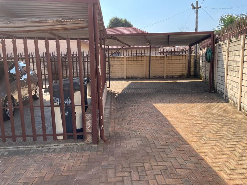 A paved outdoor carport area featuring a metal structure, enclosed by perimeter walls and security fencing.