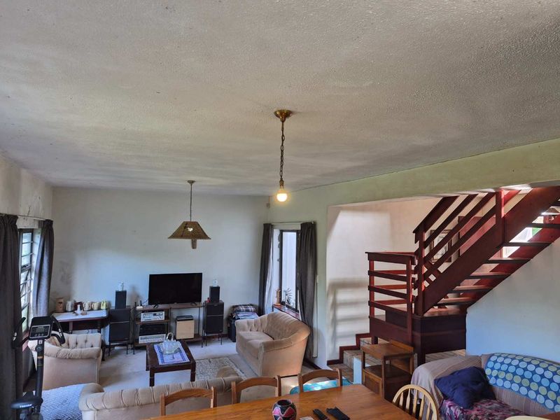 Open plan living and dining area featuring a prominent wooden staircase, large windows, and original ceiling finishes in need of cosmetic updates.