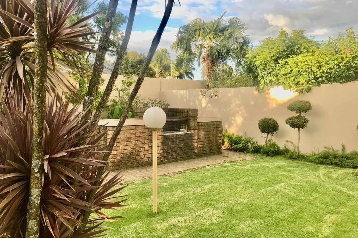 A private rear garden featuring a brick built-in braai, paved patio area, and well-maintained lawn enclosed by a perimeter wall.