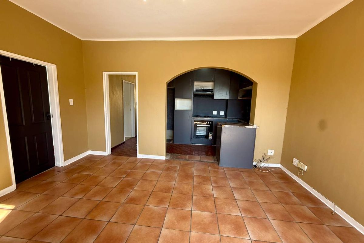 An empty open-plan living area featuring tiled flooring, an arched entryway to the kitchen, and built-in kitchen cabinetry with an integrated oven.