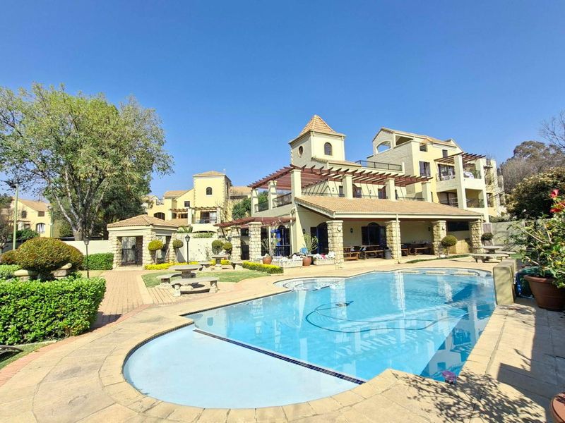 A well-maintained outdoor pool area featuring stone paving, a pergola-covered patio, and landscaped gardens within a residential apartment complex.