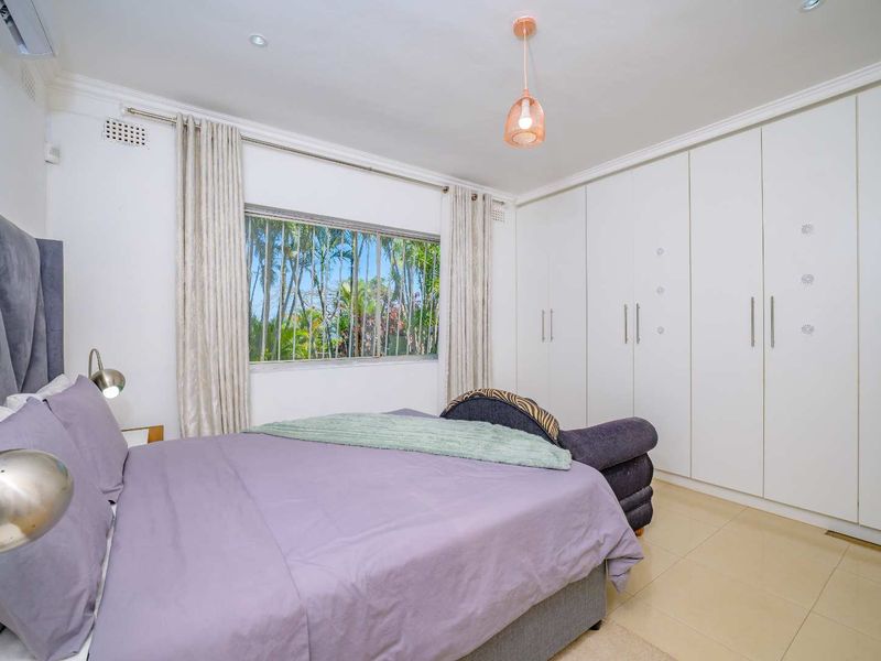 A well-maintained bedroom featuring extensive built-in cabinetry, tiled flooring, air conditioning, and large windows with security bars overlooking a lush garden.