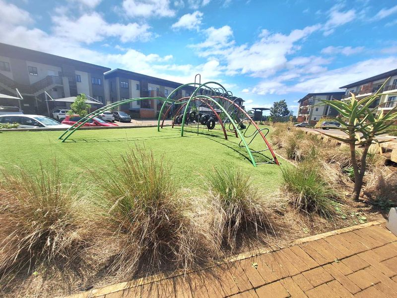 A well-maintained communal garden area featuring artificial turf, landscaped beds, and a playground structure within an apartment complex.