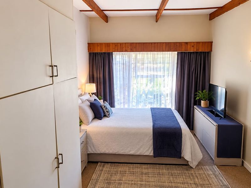 A cozy bedroom featuring exposed timber ceiling beams, built-in cupboards, and a large window with curtains, providing a comfortable and functional living space.