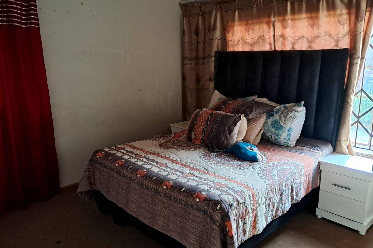 A bedroom featuring a window with security bars and curtains, showing signs of wear on the walls and flooring.