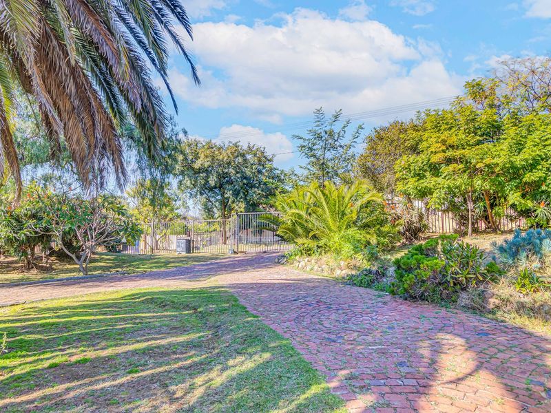 A view of the property entrance featuring a cobblestone driveway leading to a metal security gate, surrounded by established trees and garden landscaping.