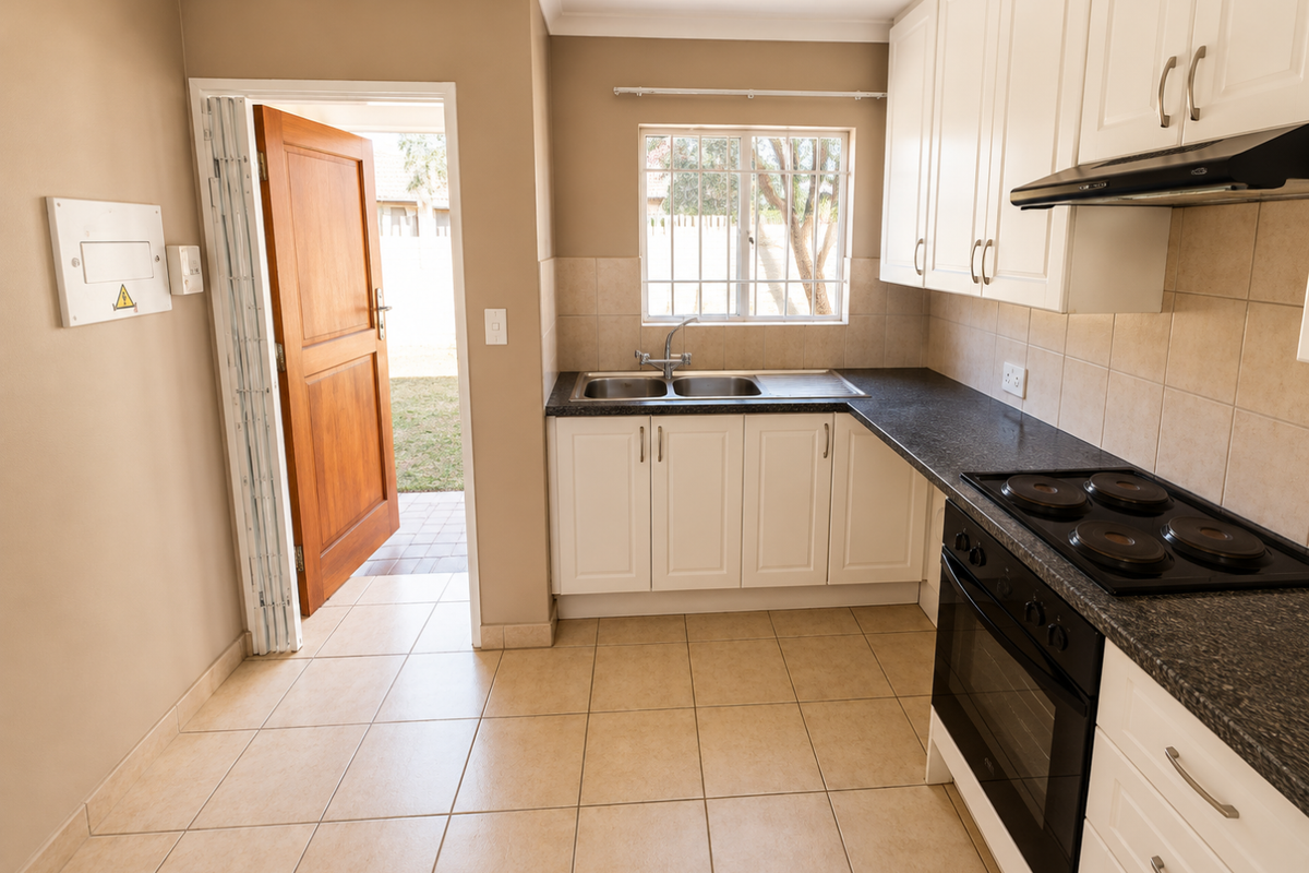 A clean, functional kitchen featuring white cabinetry, dark countertops, built-in oven and hob, tiled flooring, and direct access to the rear exterior.
