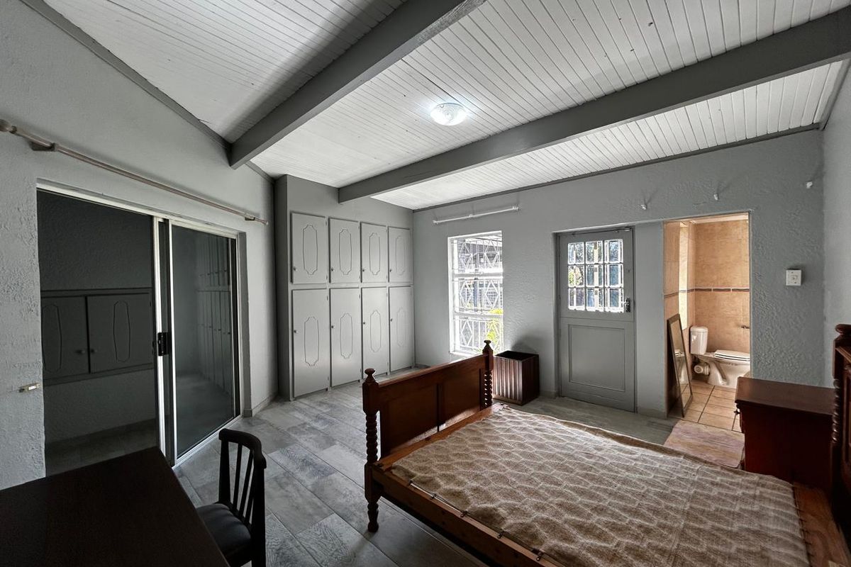 Spacious bedroom featuring exposed beam ceilings, built-in cupboards, tiled flooring, and an en-suite bathroom with direct access to an outdoor area via sliding doors.