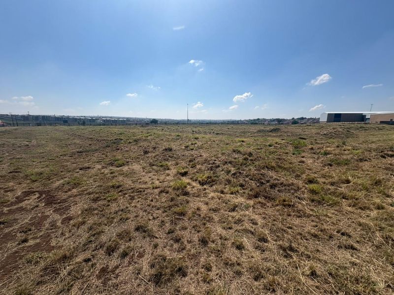 A wide-angle view of a large, vacant commercial plot of land with natural grass cover and distant industrial structures.