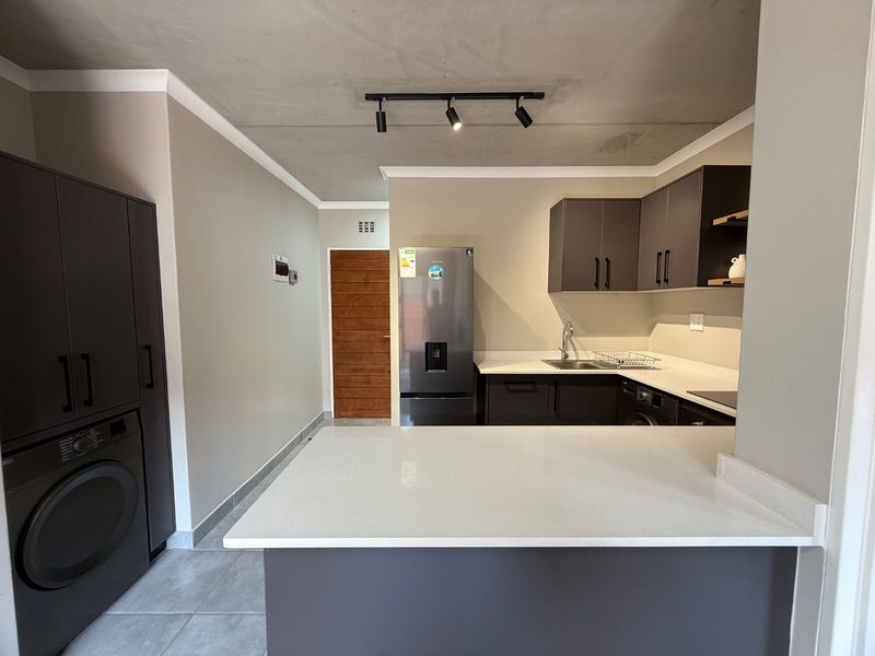 Modern kitchen featuring sleek dark cabinetry, white quartz countertops, integrated laundry space, and contemporary track lighting in an open-plan layout.