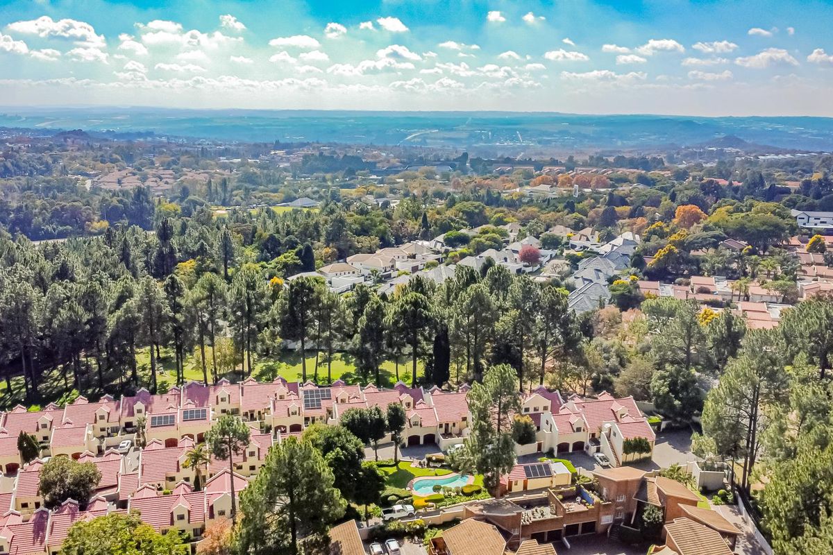 An aerial view of a townhouse complex featuring red-tiled roofs, established trees, and a communal pool area within a lush, suburban landscape.