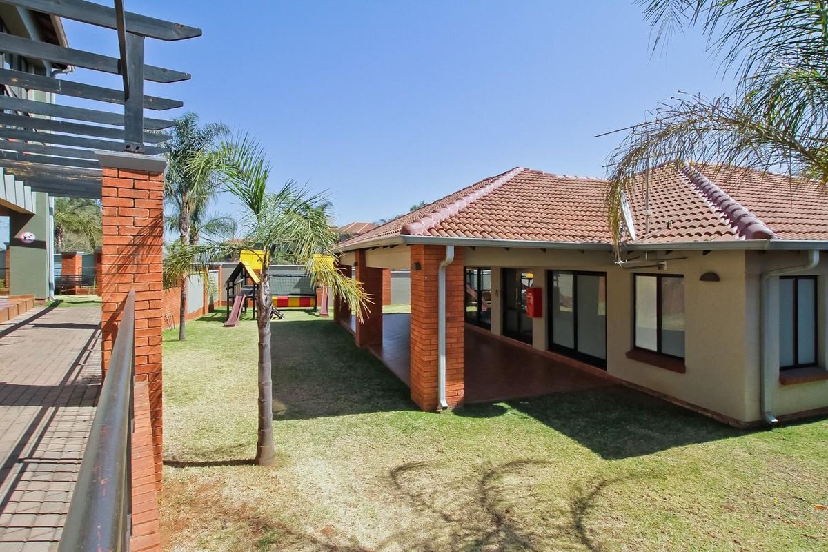 A view of the apartment complex communal area featuring a covered patio, brick pillars, tiled roof, and a landscaped garden with a playground.