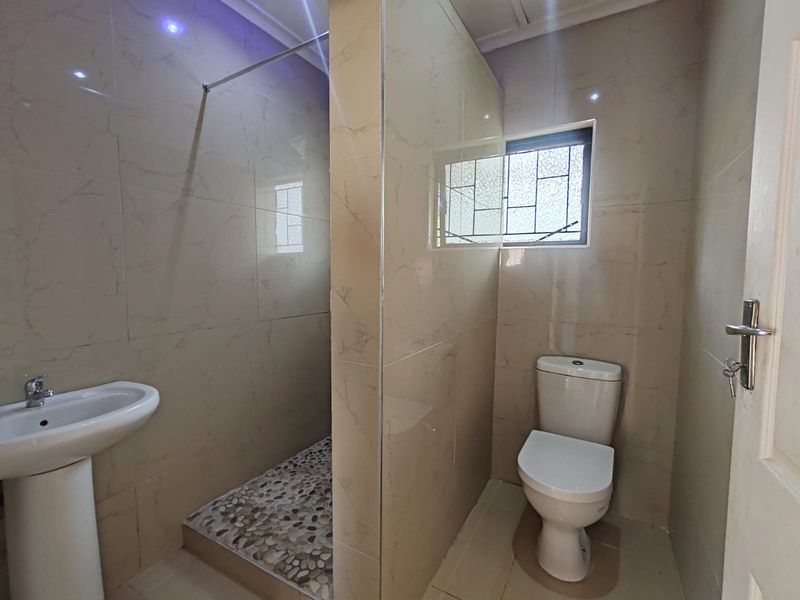 A modern, clean bathroom featuring a walk-in shower with pebble flooring, a pedestal basin, and a toilet, finished with neutral wall tiles.