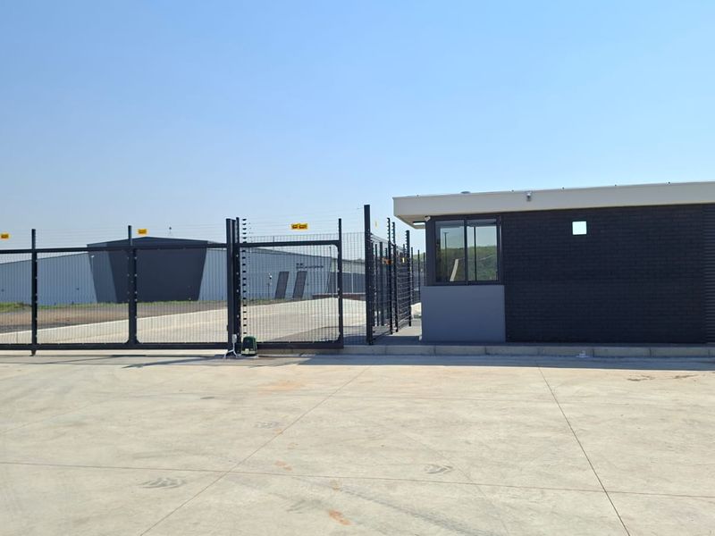 Industrial property entrance featuring a secure gate, electric fencing, and a modern guard house with a concrete driveway.