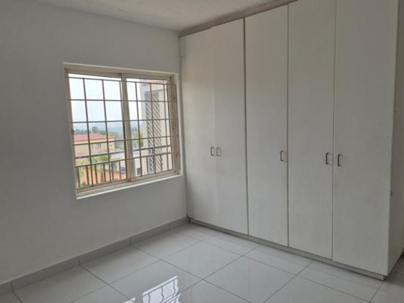 A bright, empty bedroom featuring neutral tiled flooring and ample built-in cupboard storage, complemented by a window with security bars.