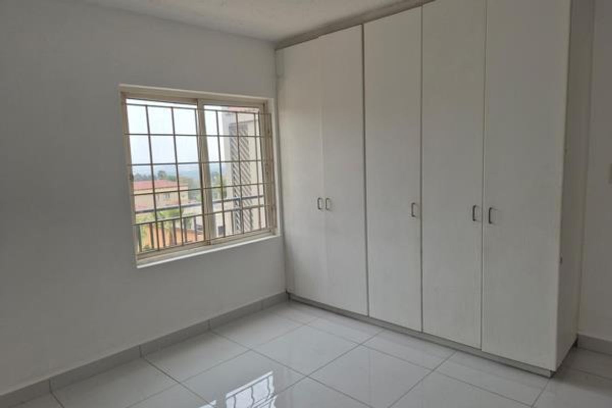 A bright, empty bedroom featuring neutral tiled flooring and ample built-in cupboard storage, complemented by a window with security bars.