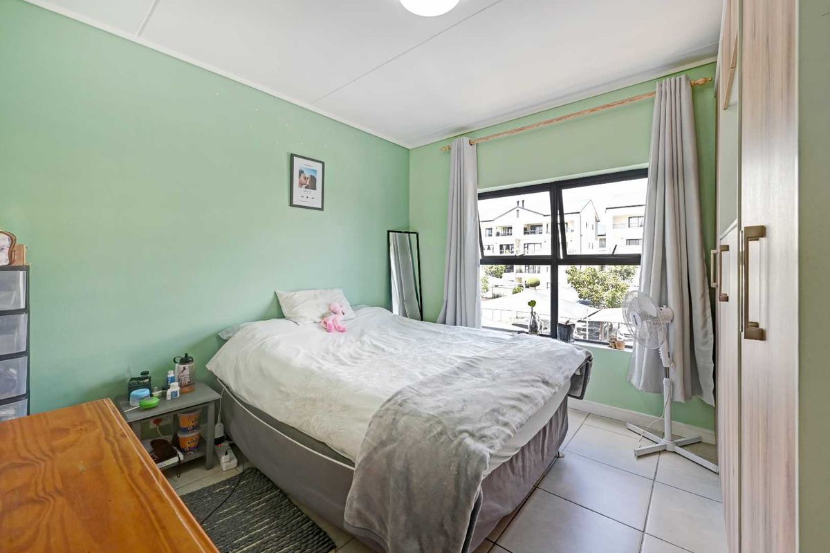 A bedroom featuring light green walls, tiled flooring, and a large window providing natural light and views of the surrounding apartment complex.