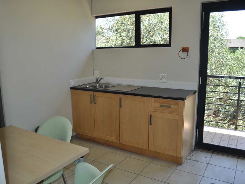A compact office kitchenette featuring light wood cabinetry, a stainless steel sink, black countertop, and direct access to an outdoor balcony area.