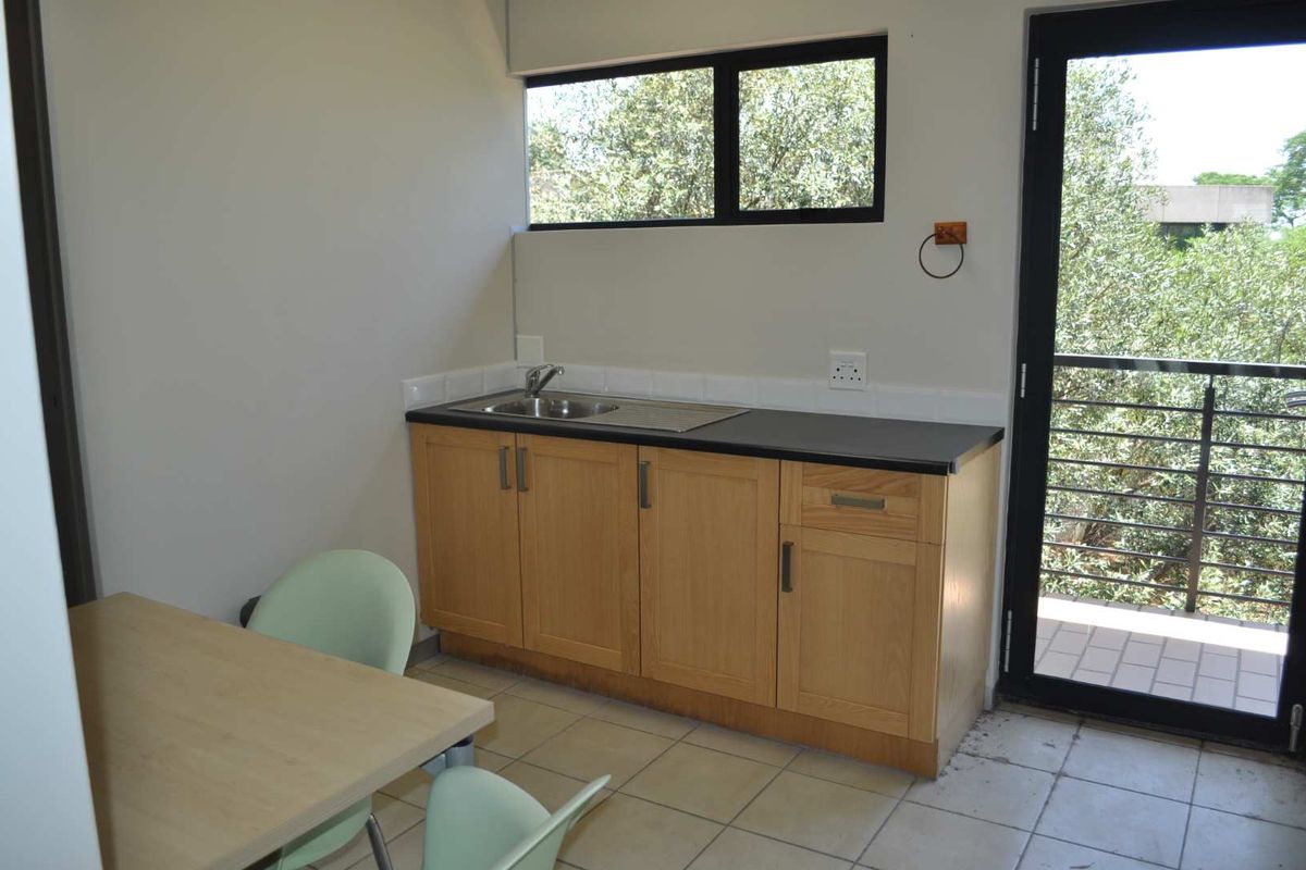 A compact office kitchenette featuring light wood cabinetry, a stainless steel sink, black countertop, and direct access to an outdoor balcony area.
