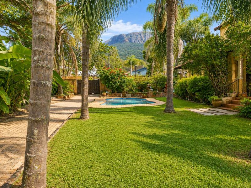 A lush, well-maintained garden featuring a swimming pool, paved driveway, and mature palm trees with a scenic mountain view in the background.