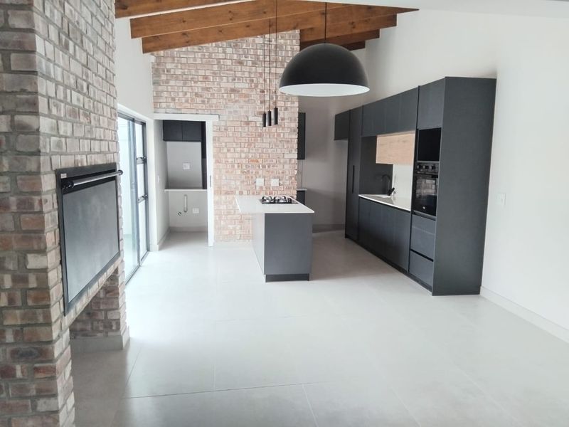 Modern open-plan living and kitchen area featuring exposed timber beams, a brick fireplace, and sleek dark cabinetry with a central island.
