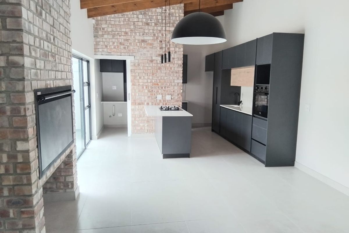 Modern open-plan living and kitchen area featuring exposed timber beams, a brick fireplace, and sleek dark cabinetry with a central island.