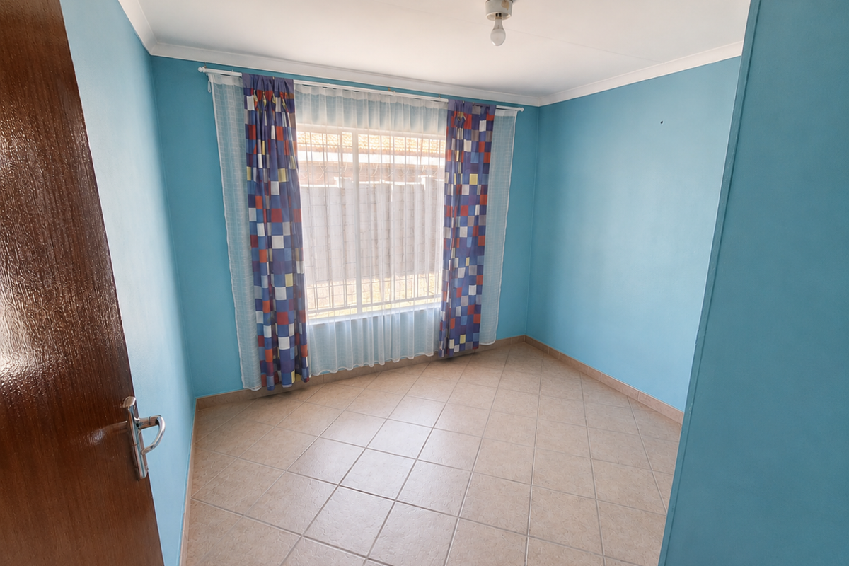 A bright, empty bedroom featuring light blue walls, tiled flooring, and a window with security bars.