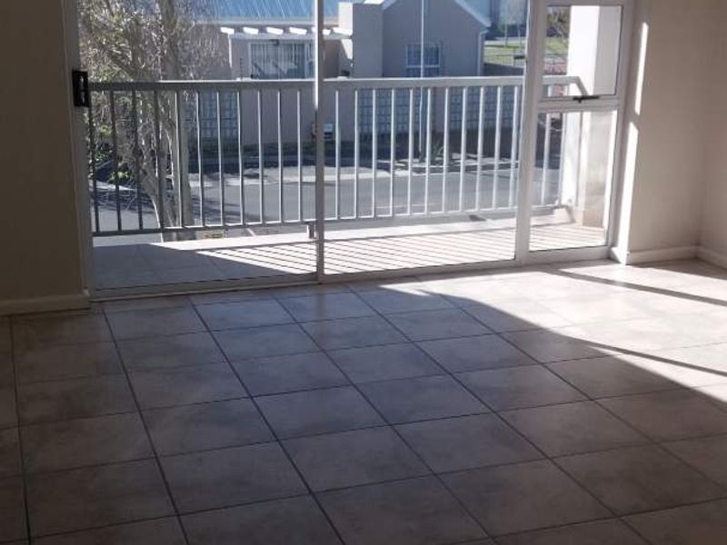 An empty, sunlit living area featuring neutral tiled flooring and a large sliding glass door providing access to a private balcony.