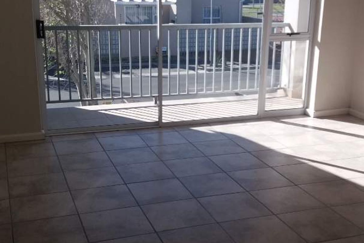 An empty, sunlit living area featuring neutral tiled flooring and a large sliding glass door providing access to a private balcony.