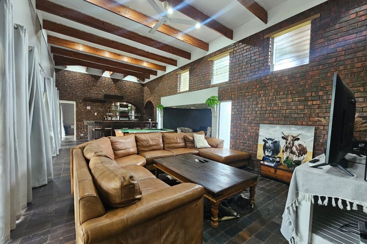 Spacious living area featuring exposed timber ceiling beams, rustic face-brick walls, and tiled flooring, with an integrated bar area in the background.