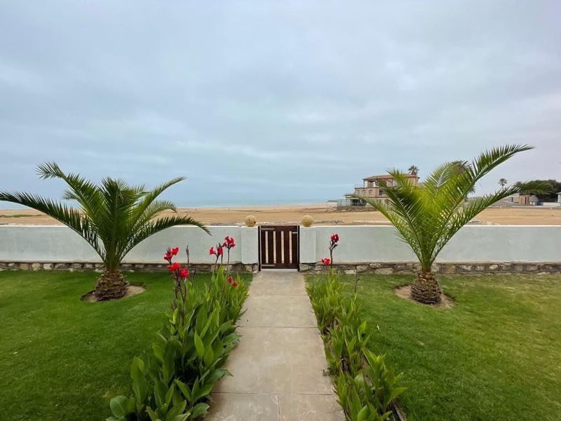 A well-maintained rear garden featuring a paved walkway, manicured lawn, and a boundary wall with a pedestrian gate leading toward the beach.