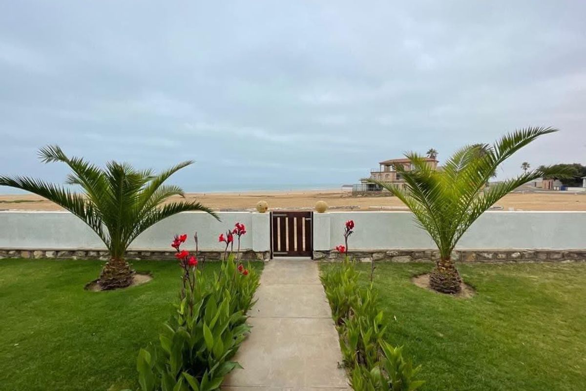 A well-maintained rear garden featuring a paved walkway, manicured lawn, and a boundary wall with a pedestrian gate leading toward the beach.
