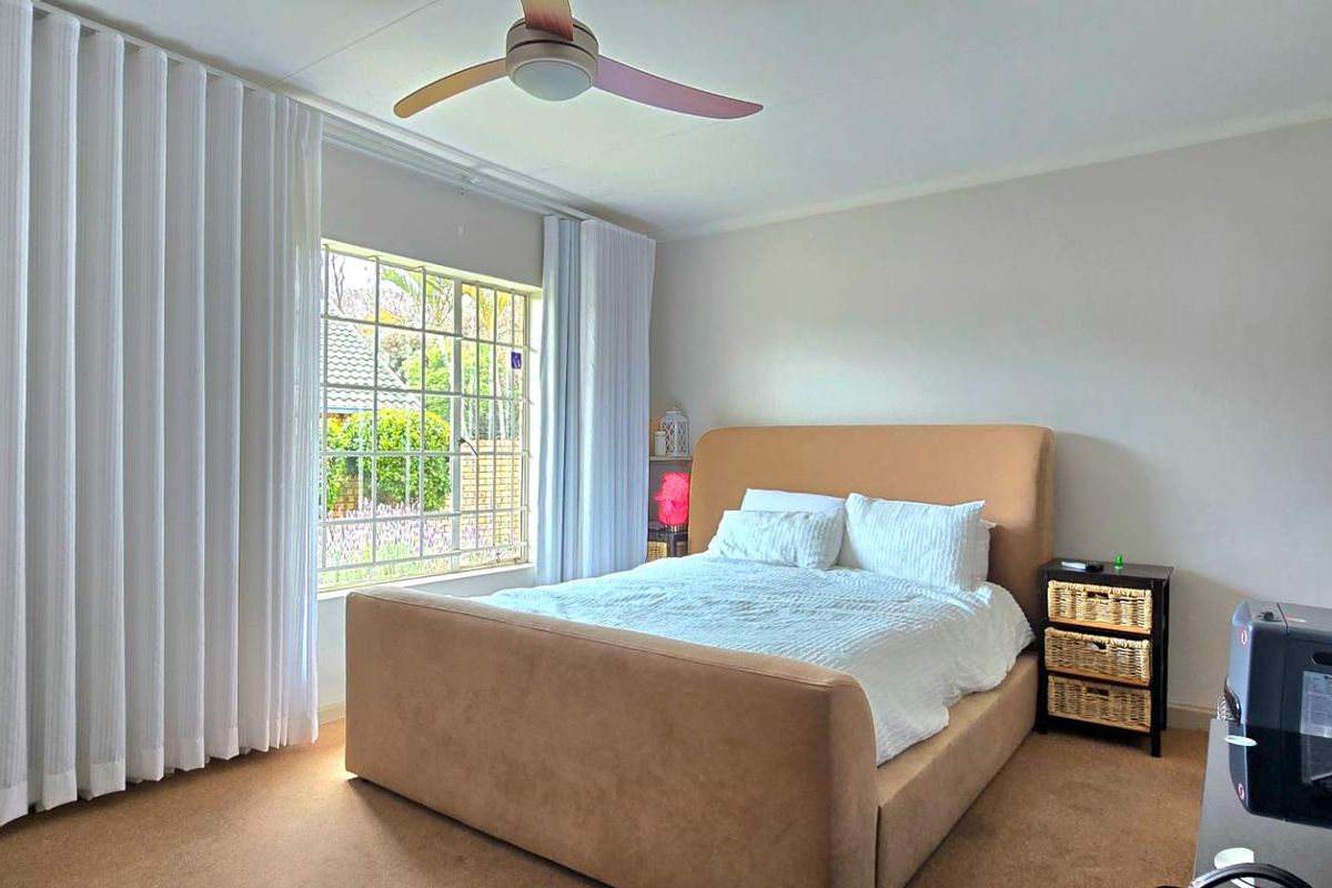 A well-lit bedroom featuring carpeted flooring, a ceiling fan, and a large window with security bars, providing a comfortable and functional living space.