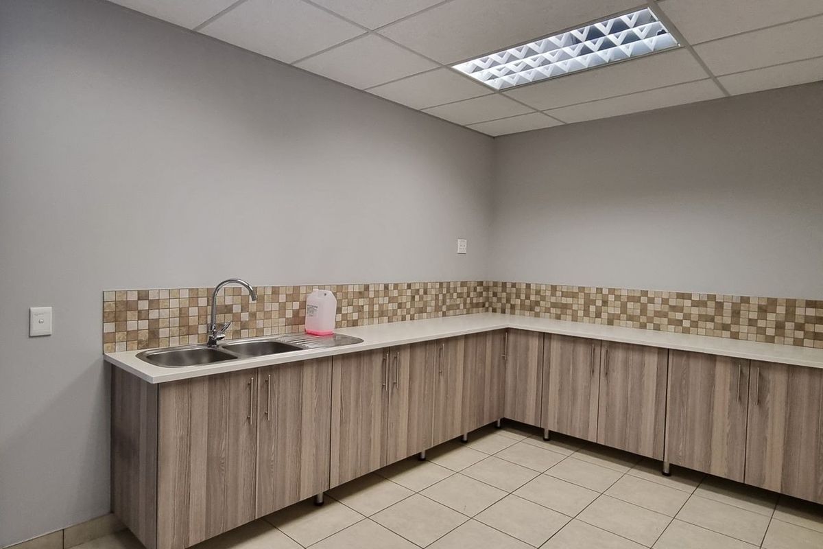 A functional office kitchenette featuring wood-look cabinetry, a double sink, tiled splashback, and recessed ceiling lighting.
