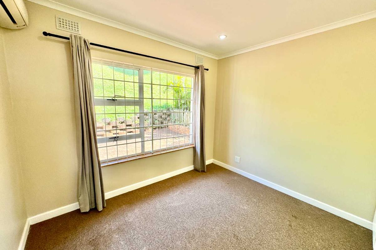 A bright, empty bedroom featuring neutral carpeting, a large window with security bars, and a wall-mounted air conditioning unit.