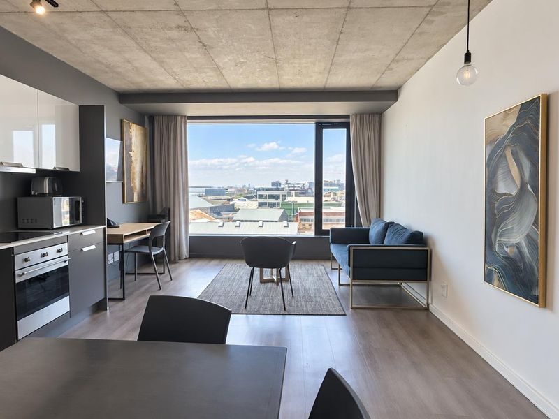 Modern open-plan apartment featuring an exposed concrete ceiling, sleek kitchen cabinetry, integrated oven, and a large window offering expansive city views.