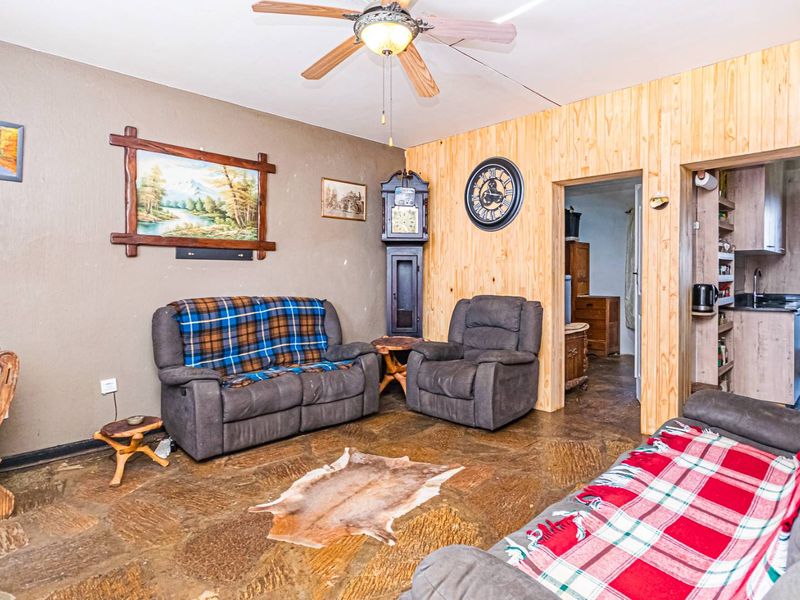 Living room featuring rustic stone flooring, wood-paneled walls, and a ceiling fan, with an adjacent doorway leading to a kitchen area.