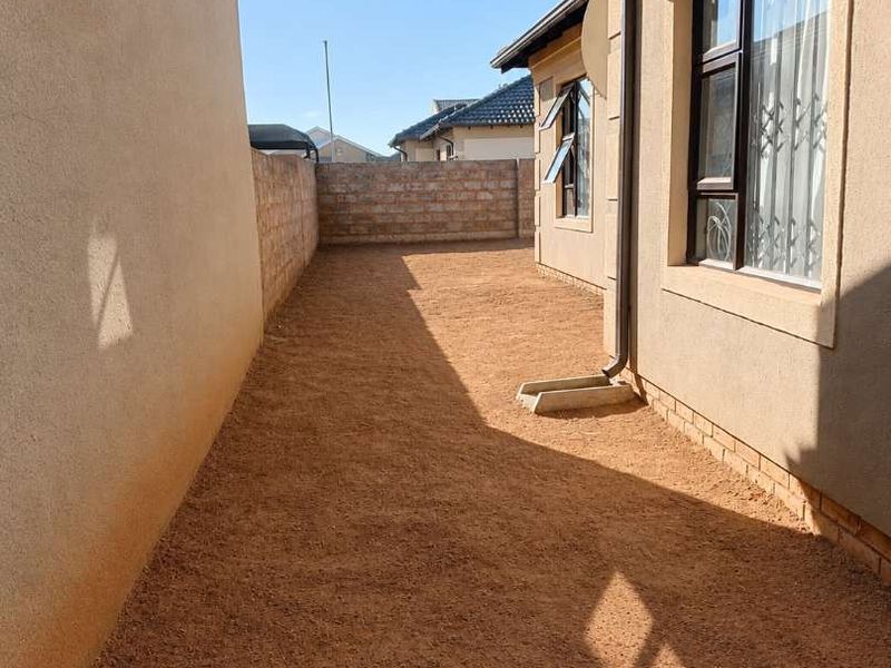 A side exterior view of the property featuring a gravel ground surface, brick boundary wall, and aluminium window frames with security bars.