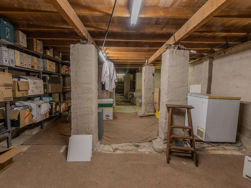A functional utility or storage basement area featuring exposed timber ceiling beams, concrete support columns, and stone flooring, suitable for workshop or storage use.