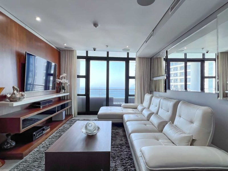 Modern living room featuring built-in wooden media cabinetry, recessed ceiling lighting, and large glass sliding doors leading to a balcony with ocean views.