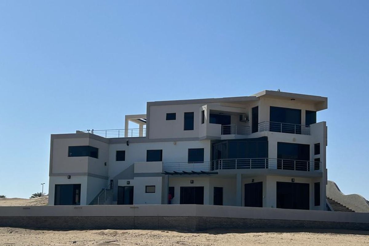 A modern multi-story coastal residence featuring multiple balconies with stainless steel railings, a flat roof design, and a perimeter retaining wall.