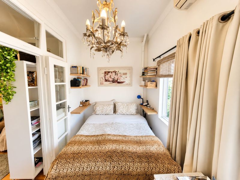 A compact bedroom featuring built-in shelving, a classic chandelier, and a window with integrated air conditioning unit.