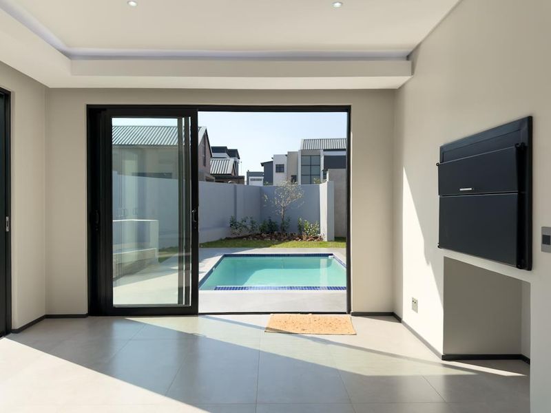 Modern living area featuring large sliding doors opening to a private pool and garden, complete with a built-in braai and neutral tiled flooring.