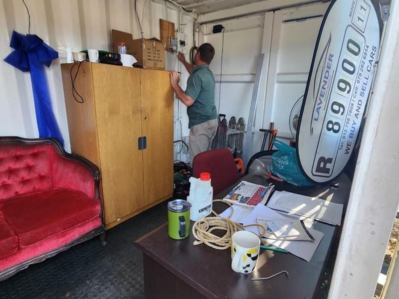 A functional office space housed within a converted shipping container, featuring exposed electrical infrastructure and durable rubber flooring.