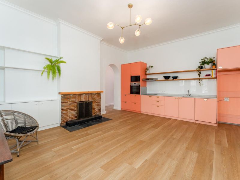 Open-plan living and kitchen area featuring modern peach-toned cabinetry, integrated appliances, a classic brick fireplace, and light timber flooring.