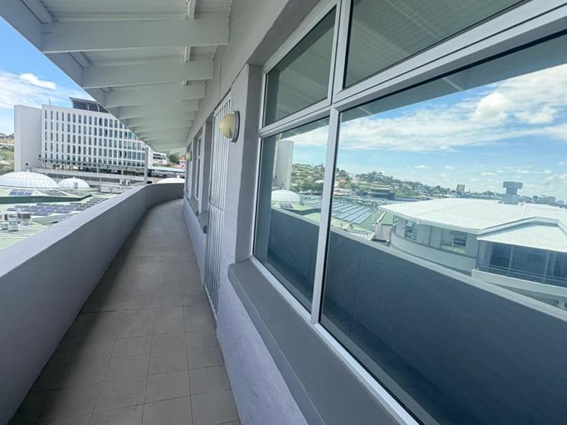 An exterior covered balcony walkway featuring tiled flooring, aluminum-framed windows, and a security gate, offering an elevated view of the surrounding office complex.