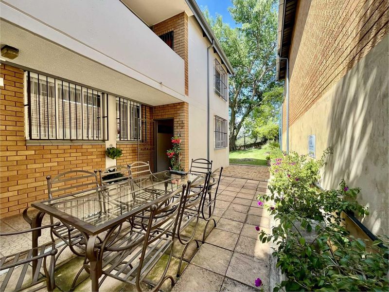 A paved rear patio area featuring brick exterior walls, security bars on windows, and access to a shared garden space.