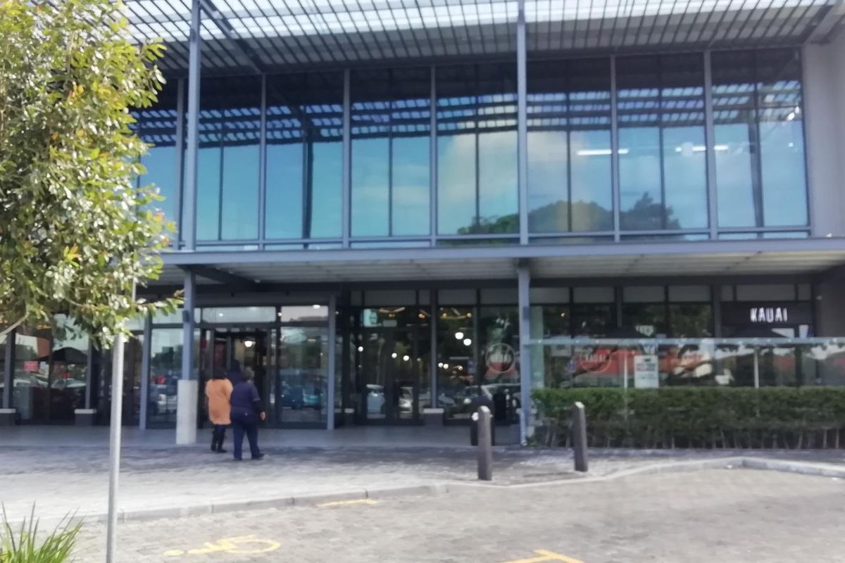 Modern retail building facade featuring extensive glass curtain walling, steel structural elements, and a paved pedestrian entrance area.