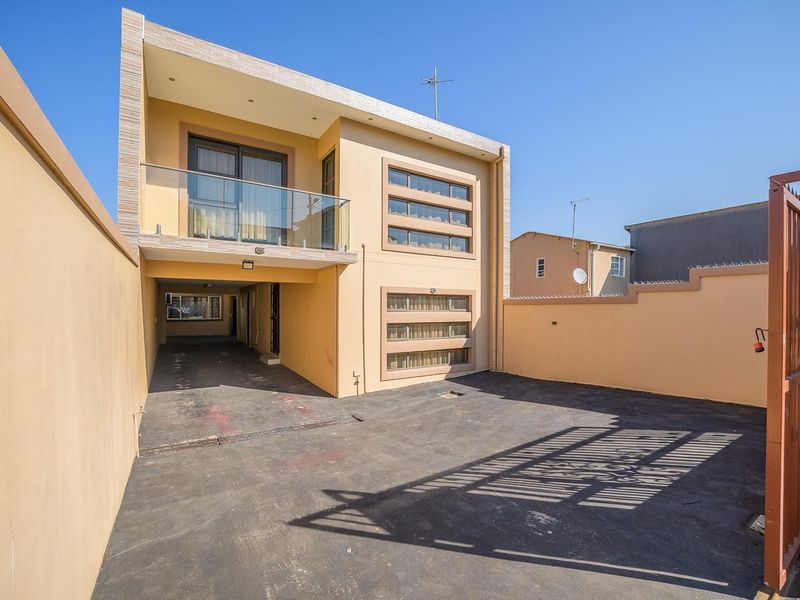 Front exterior view of a modern two-story house featuring a paved driveway, secure perimeter wall, and a balcony with a glass balustrade.